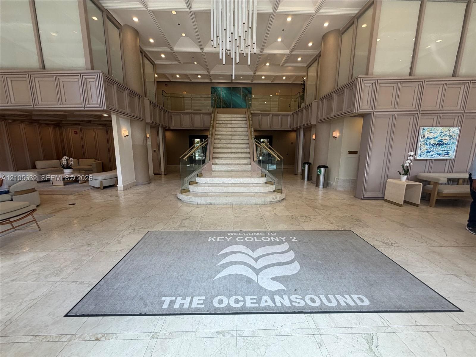 Grand lobby featuring a coffered ceiling, stone flooring, a multi-tier contemporary chandelier, a central stone staircase with glass railing, and decorative wall paneling