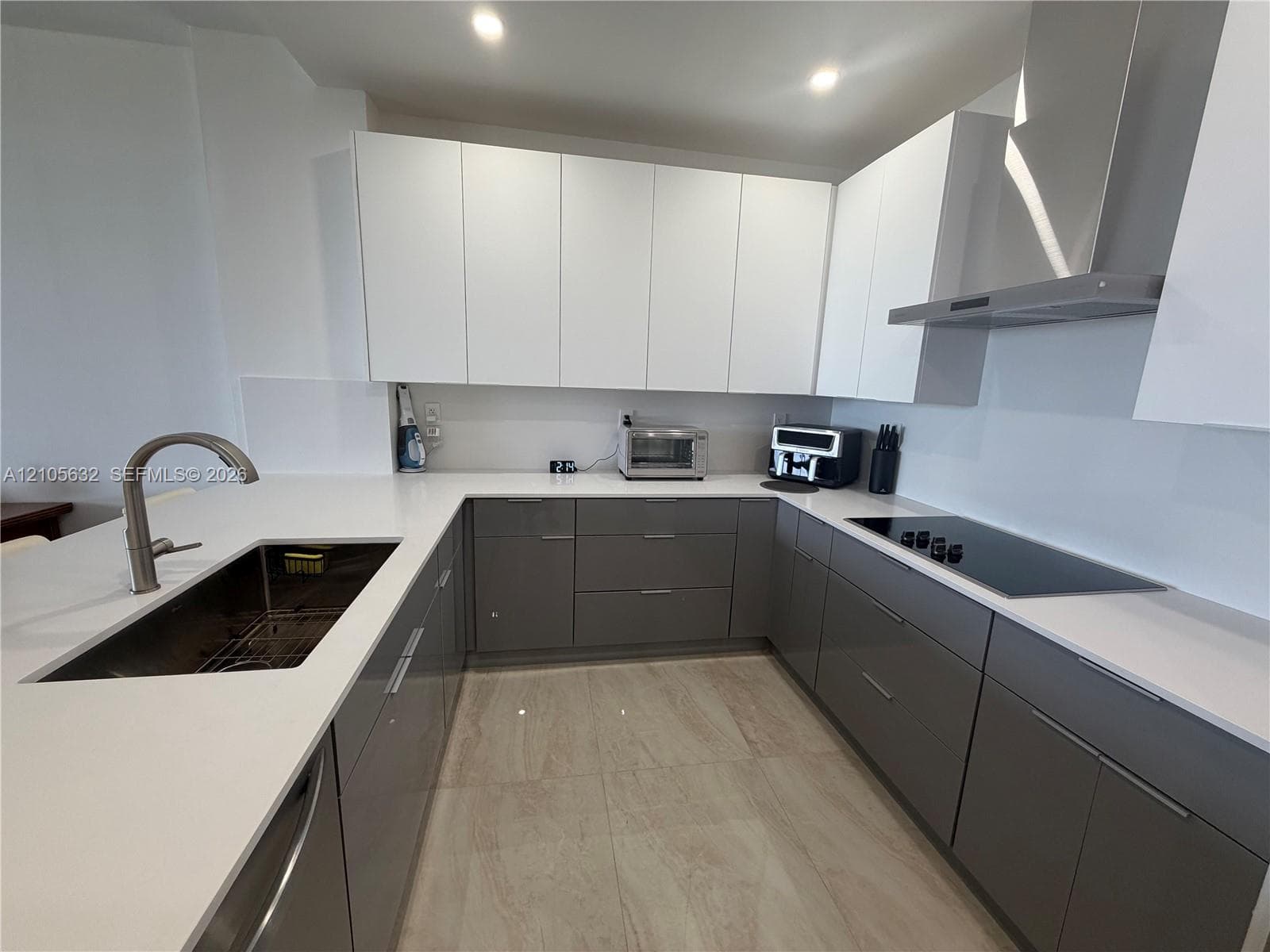 Contemporary kitchen featuring two-tone flat-panel cabinetry, light-toned solid surface countertops, a stainless steel undermount sink with a gooseneck faucet, a flush-mount induction cooktop, and large-format tile flooring