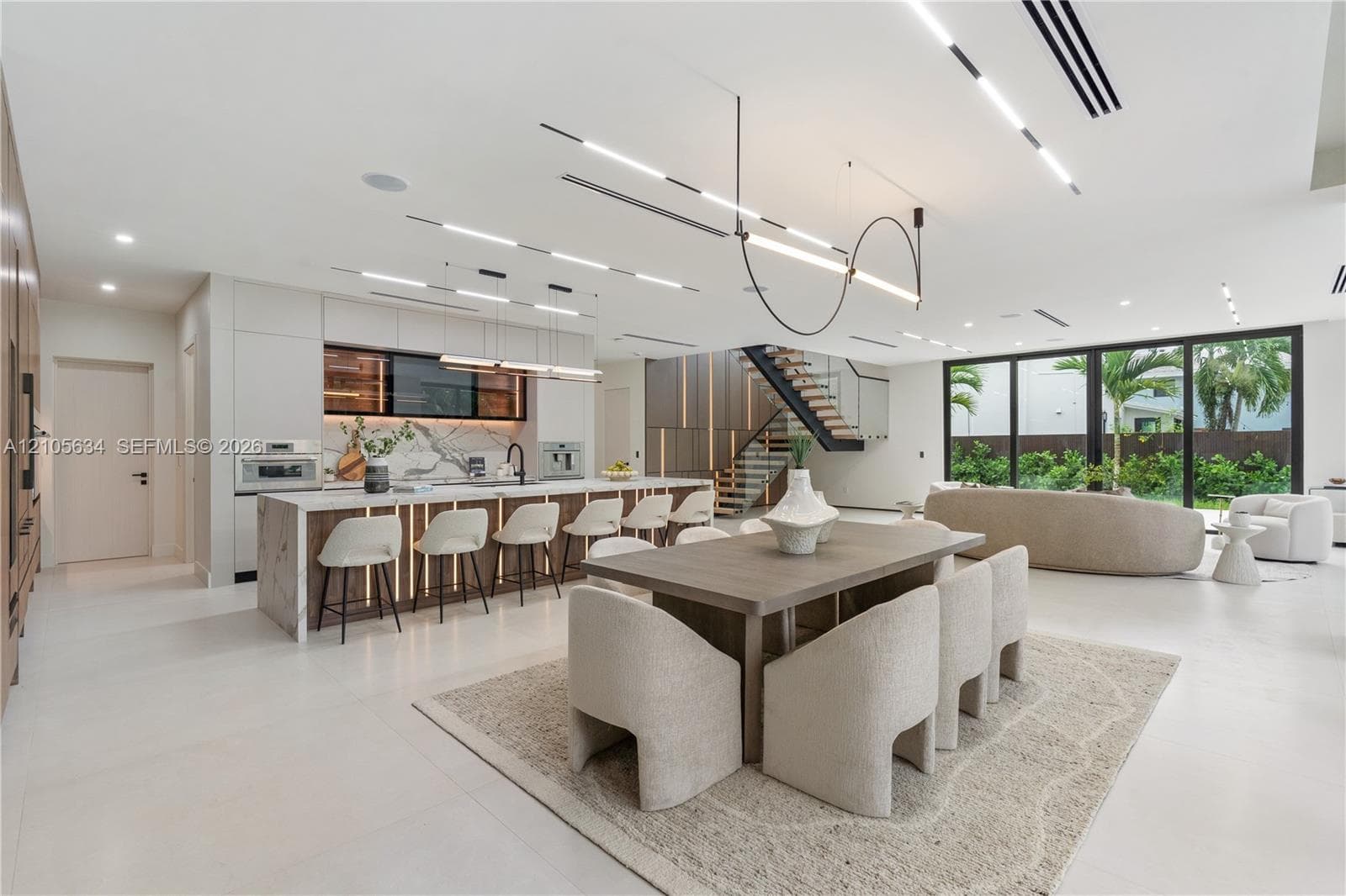 Open-concept interior featuring large-format flooring, a kitchen island with stone-surface counters, integrated LED accent lighting, floating staircase with metal railings, and floor-to-ceiling glass panel walls