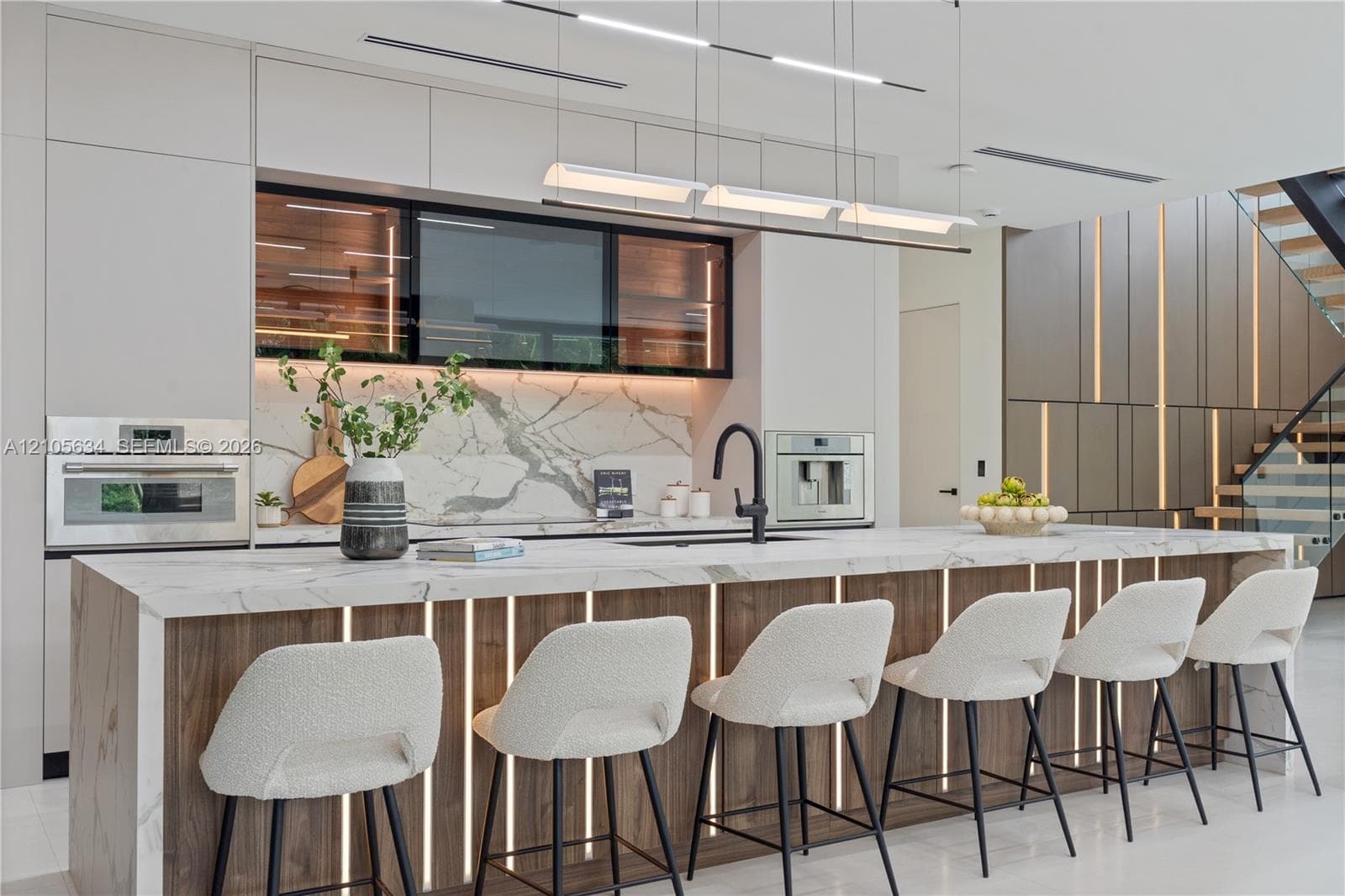 Contemporary kitchen featuring a stone-surface waterfall island with integrated vertical lighting, flat-panel cabinetry, a stone backsplash, built-in stainless steel appliances, and linear pendant lighting