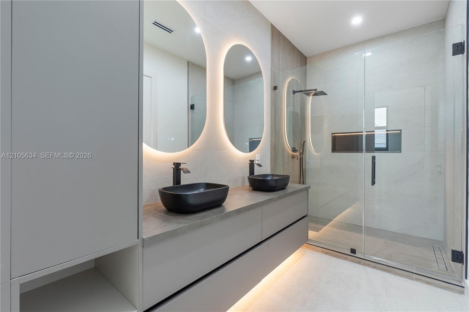 Floating dual-sink vanity with matte black fixtures, quartz-finish countertop, backlit oval mirrors, and a walk-in glass-enclosed shower featuring a built-in recessed niche