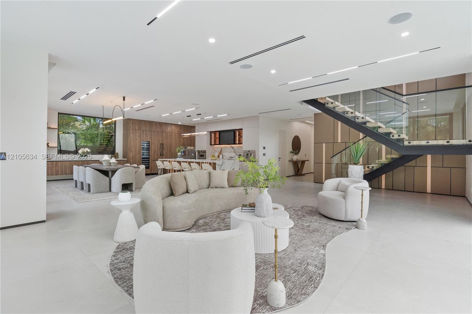 Open-concept floor plan featuring expansive stone-tile flooring, recessed linear lighting, custom wood-paneled cabinetry, and a floating staircase with glass railings