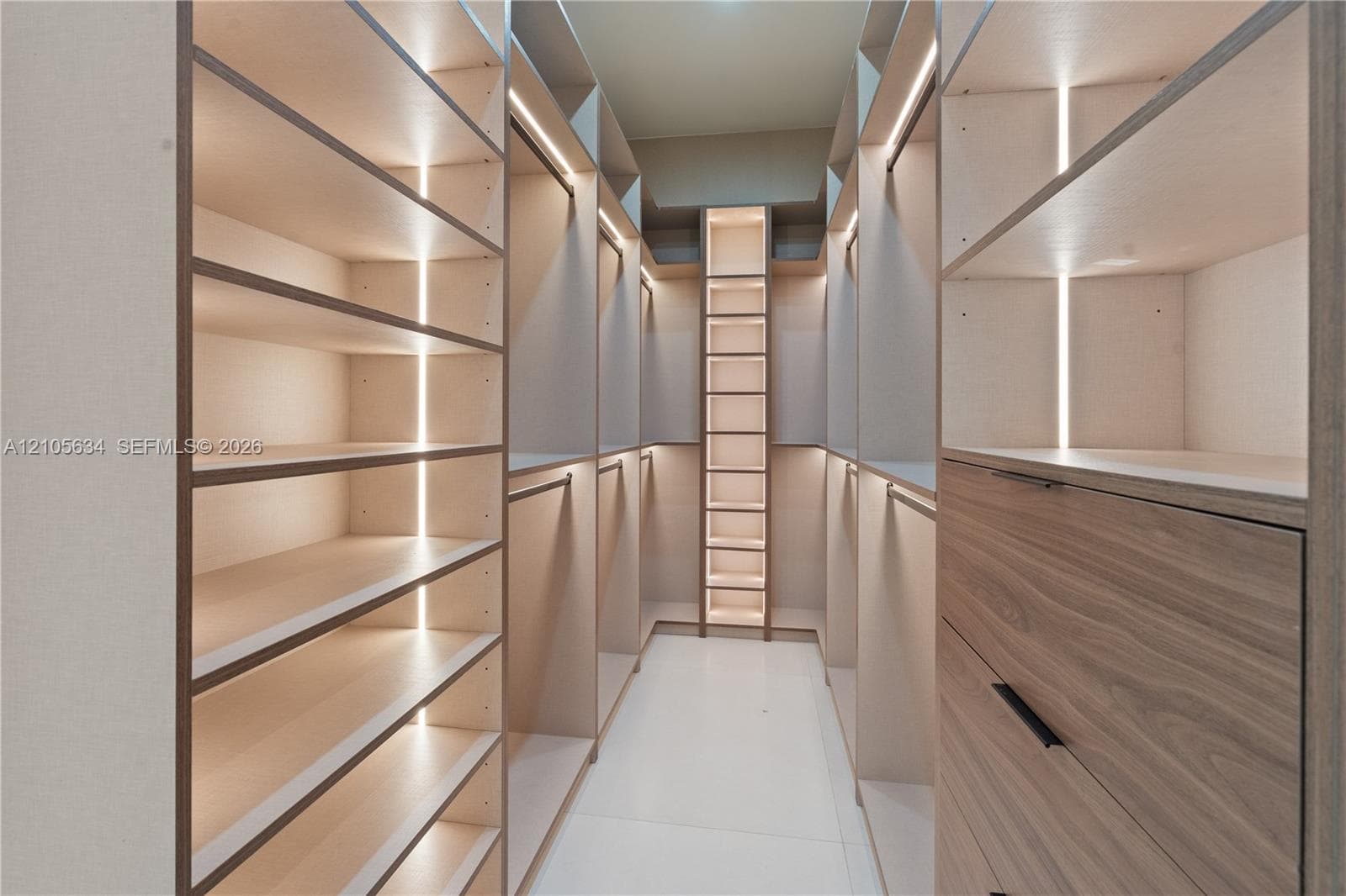 Walk-in wardrobe featuring custom cabinetry with wood-finish drawer fronts, open shelving, integrated vertical LED lighting, and matte black hardware