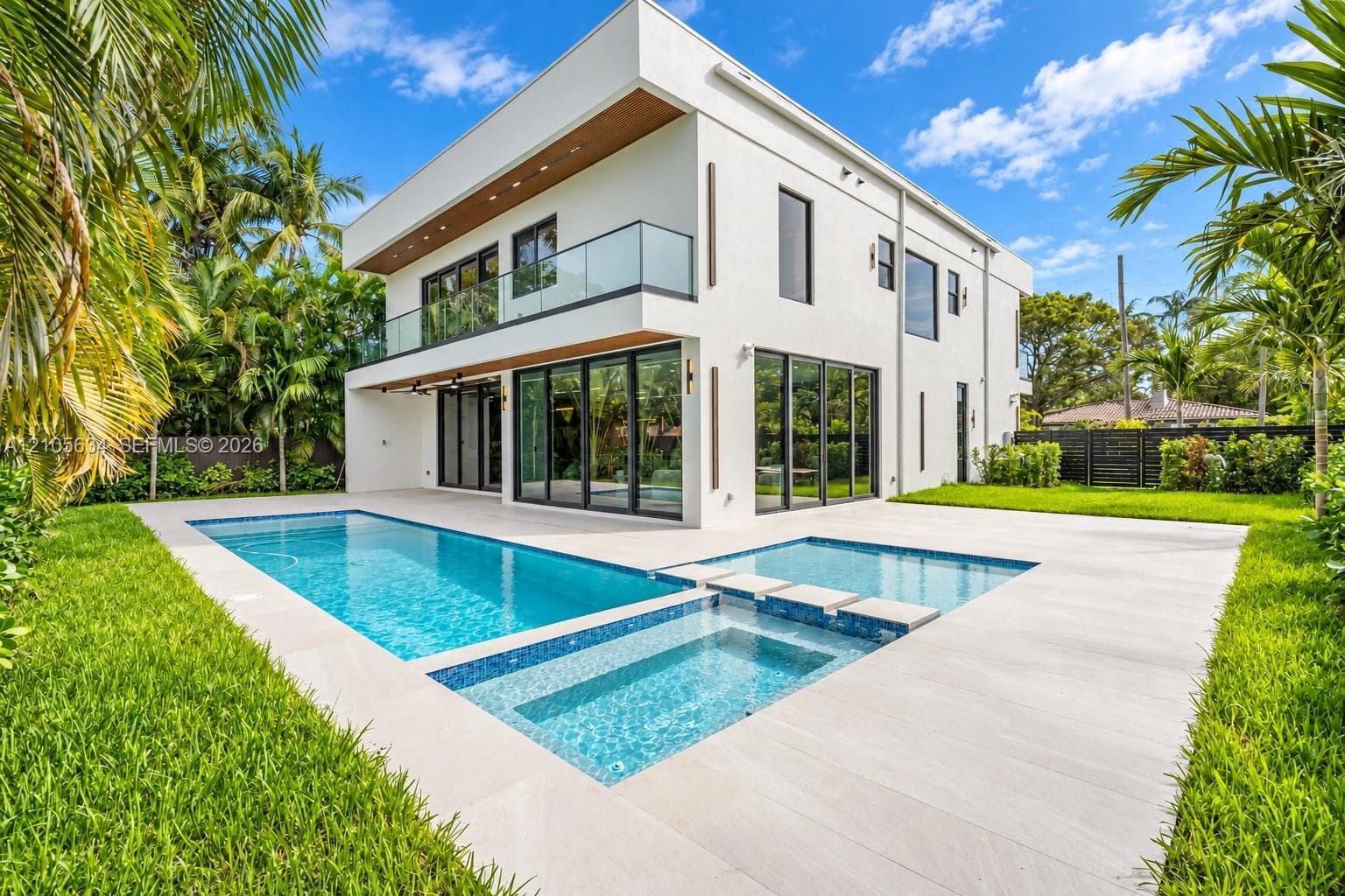 Modern two-story architectural exterior featuring smooth stucco finishes, floor-to-ceiling glass panel doors, a second-level glass-railed balcony, and an integrated swimming pool and spa surrounded by light-toned hardscaping