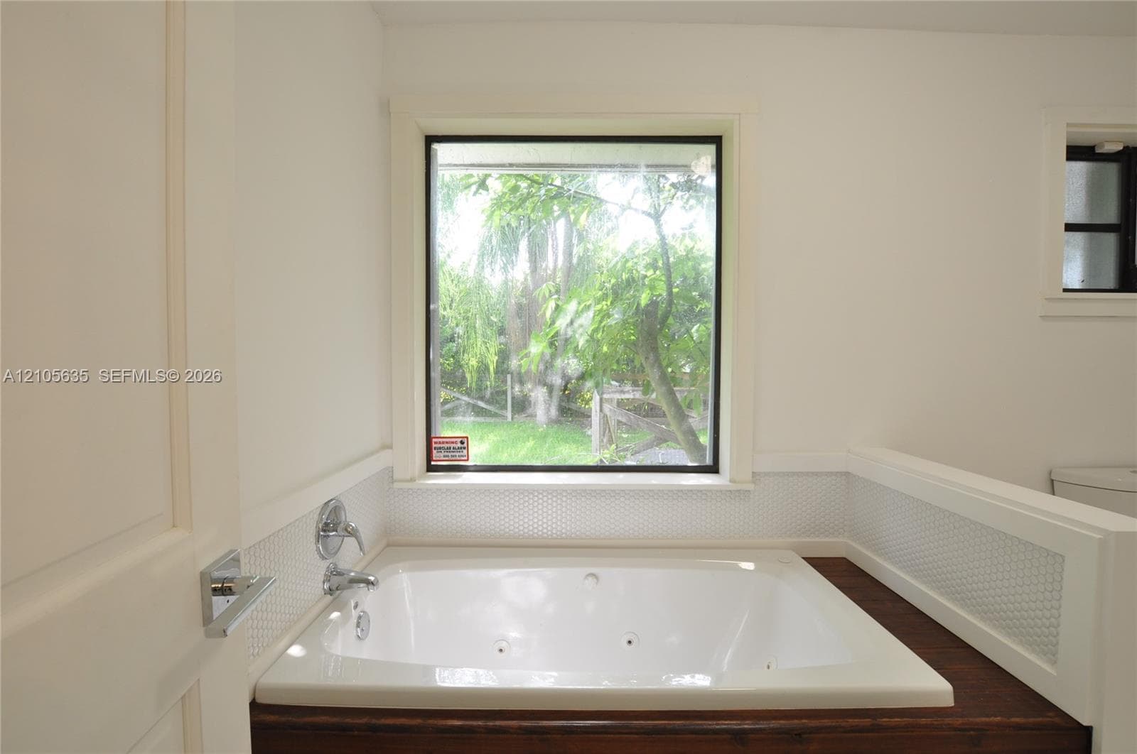 Jetted soaking tub featuring a wood-finish surround, white penny tile wainscoting, and a large exterior window