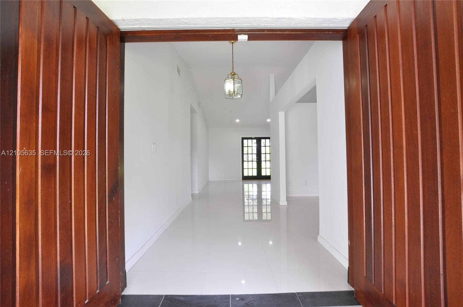 Double wood-paneled entry doors featuring polished tile flooring, white walls, and a decorative pendant light fixture