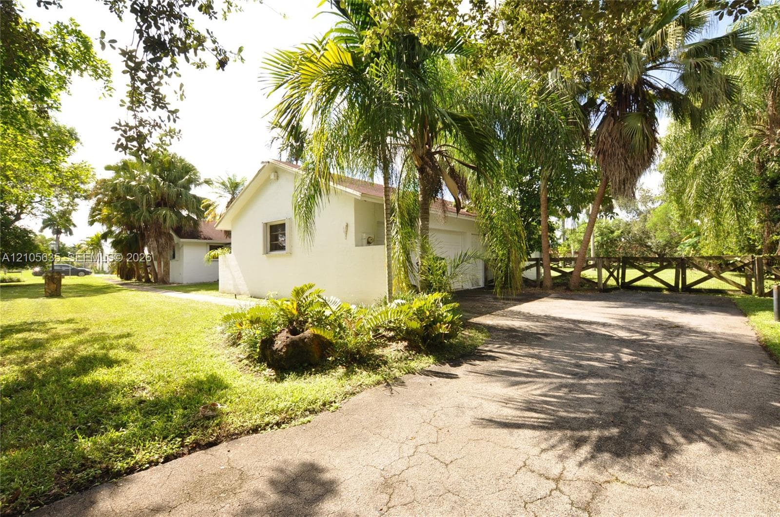 Stucco exterior, multi-car garage, asphalt driveway, timber cross-buck fencing, and established tropical landscaping
