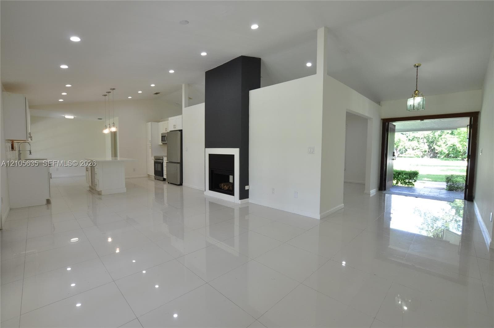 Open-concept interior featuring large-format polished tile flooring, vaulted ceilings with recessed lighting, a modern floor-to-ceiling fireplace with high-contrast surround, and stainless steel kitchen appliances