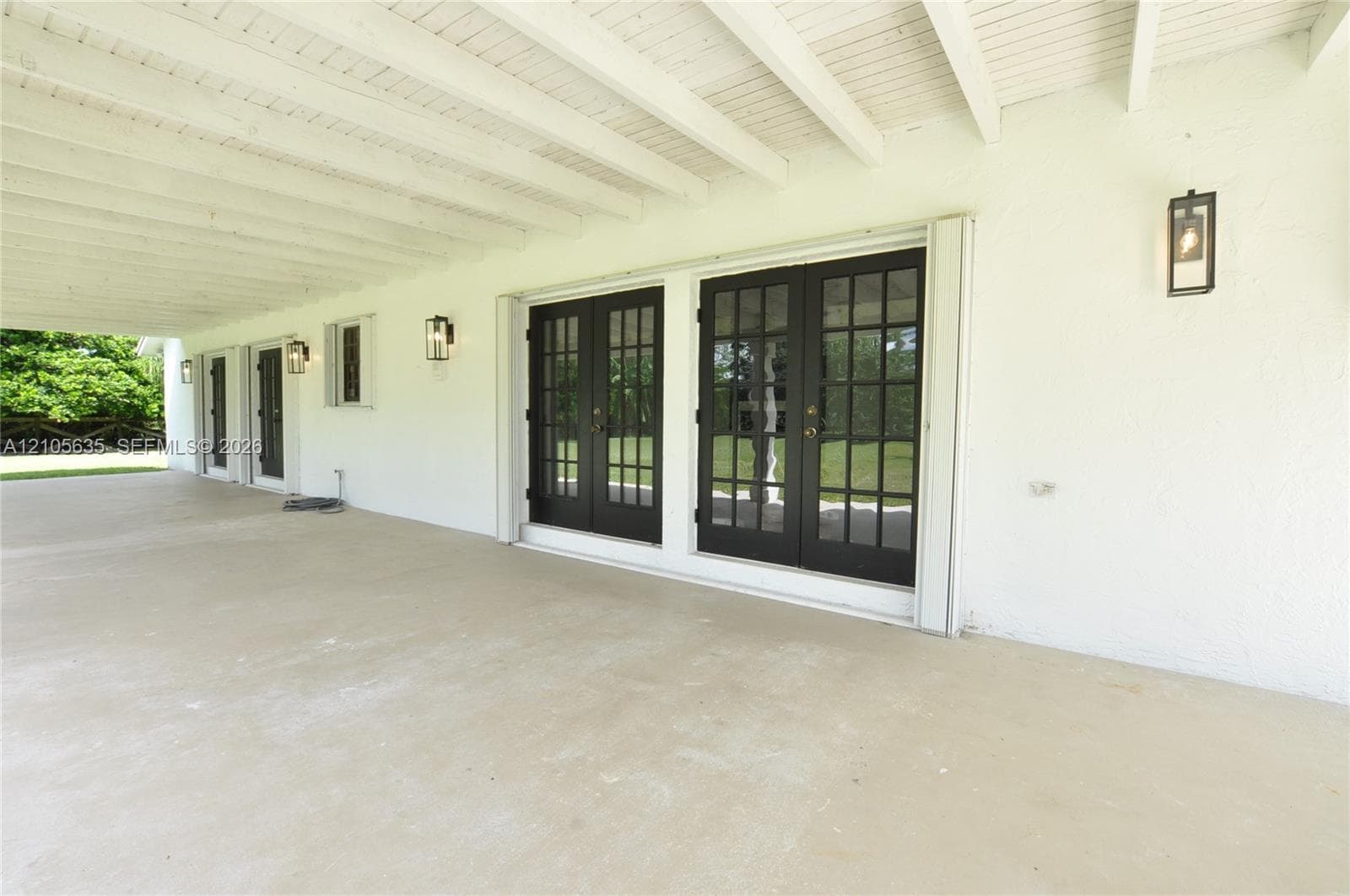 Covered patio featuring concrete flooring, white textured wall finish, exposed beam ceiling, two sets of multi-pane French doors with dark frames, and wall-mounted exterior lanterns