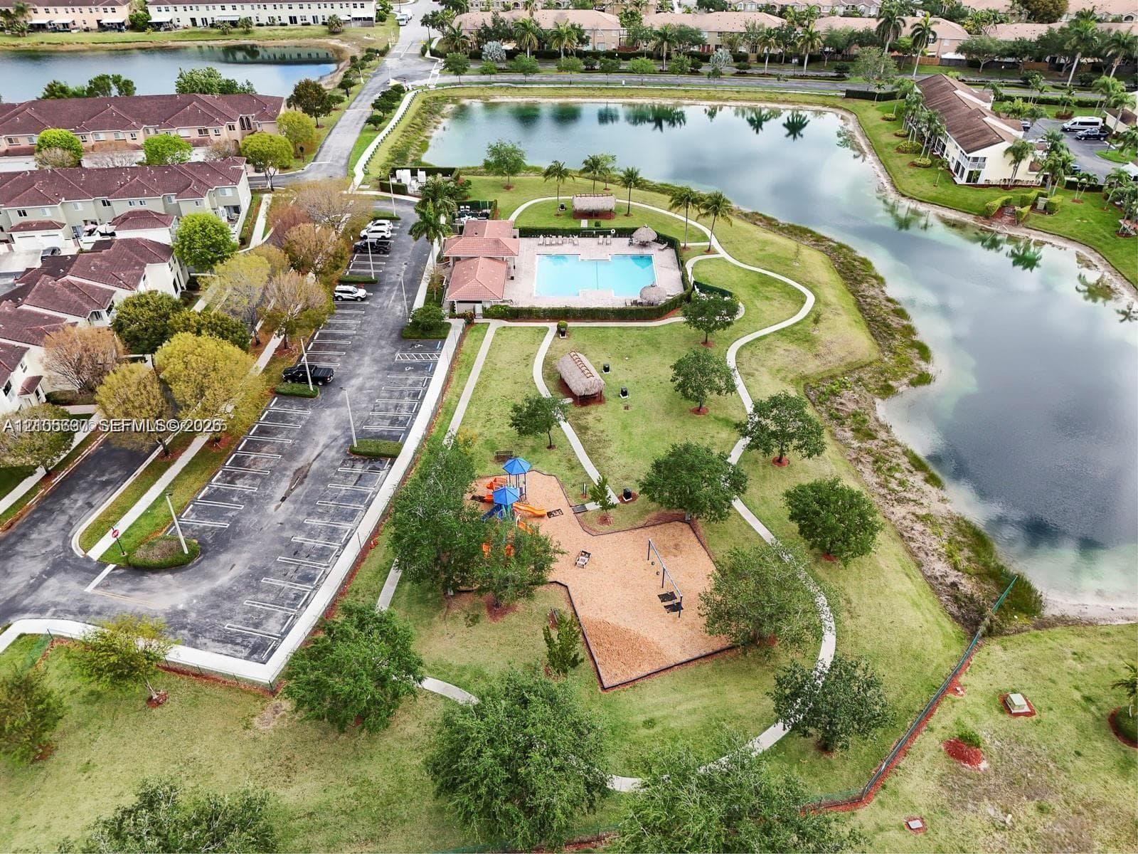 Aerial perspective of community grounds featuring a swimming pool, winding concrete pathways, a playground with integrated structures, and a large lake