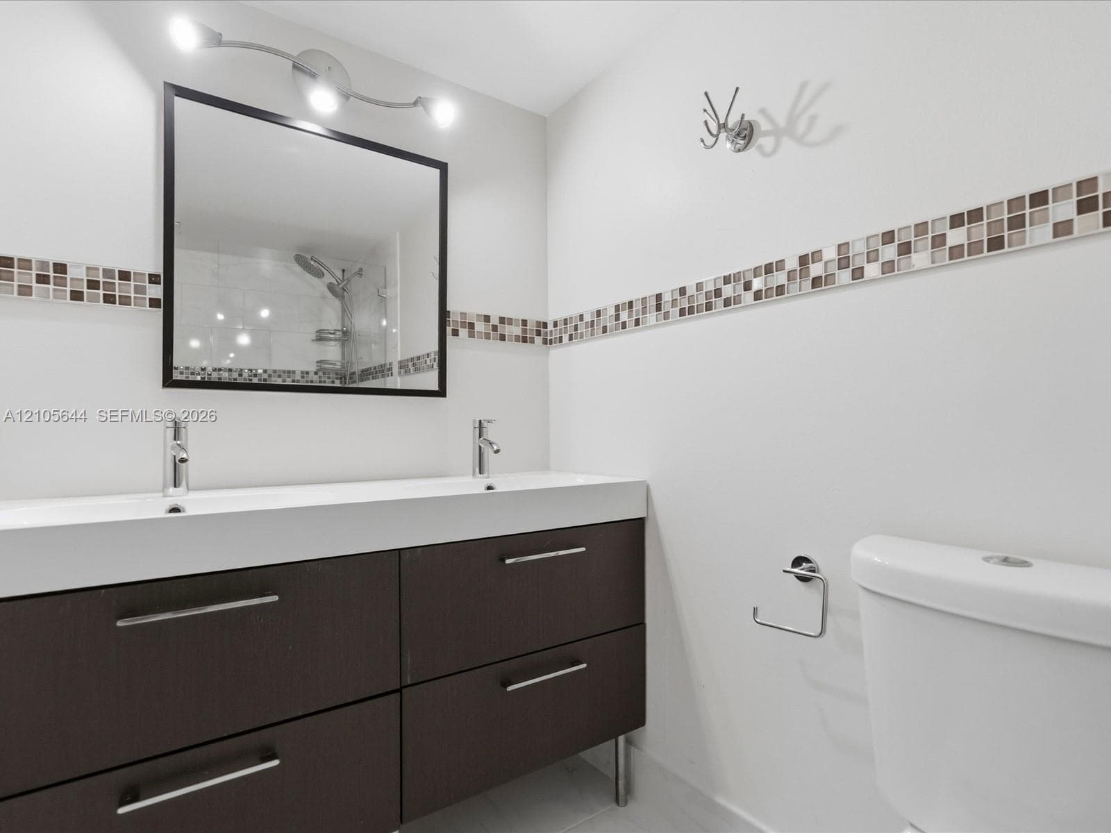 Double vanity featuring a dark-toned cabinet base with metallic hardware, integrated dual sinks, a framed mirror, and decorative mosaic tile backsplash