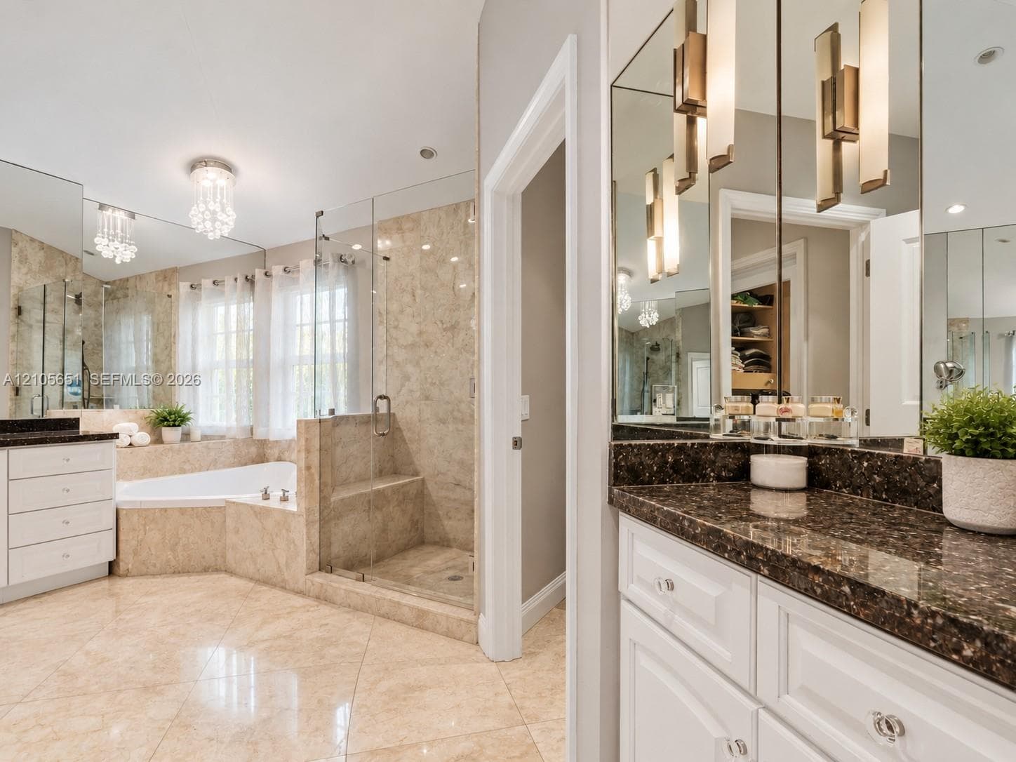 Bathroom featuring stone-finish flooring and wall tiling, dual white vanities with dark stone countertops, a frameless glass-enclosed shower with a built-in bench, a corner soaking tub, and custom recessed and pendant lighting