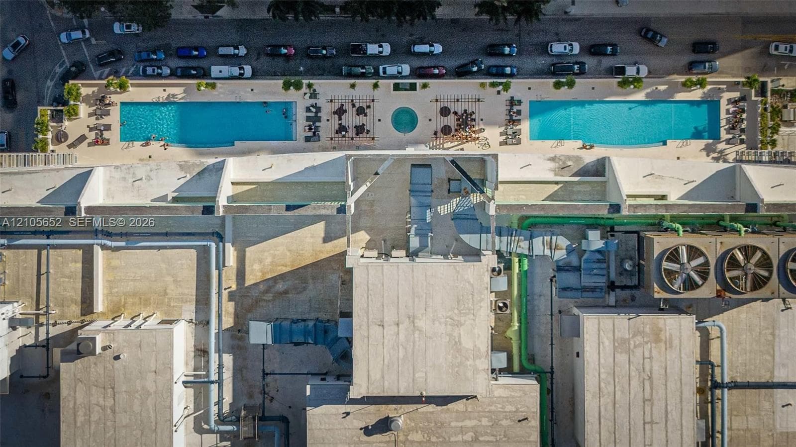 Aerial perspective of property amenities featuring two rectangular swimming pools, a central circular spa, and paved sundeck areas with pergola structures
