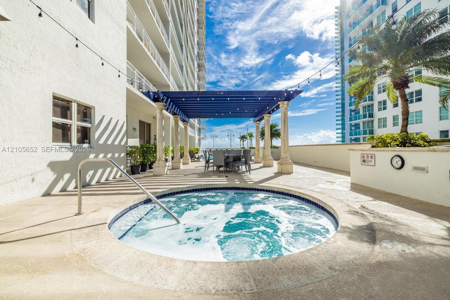 In-ground circular spa featuring a stainless steel handrail, shaded pergola with decorative stone columns, and overhead bistro-style lighting