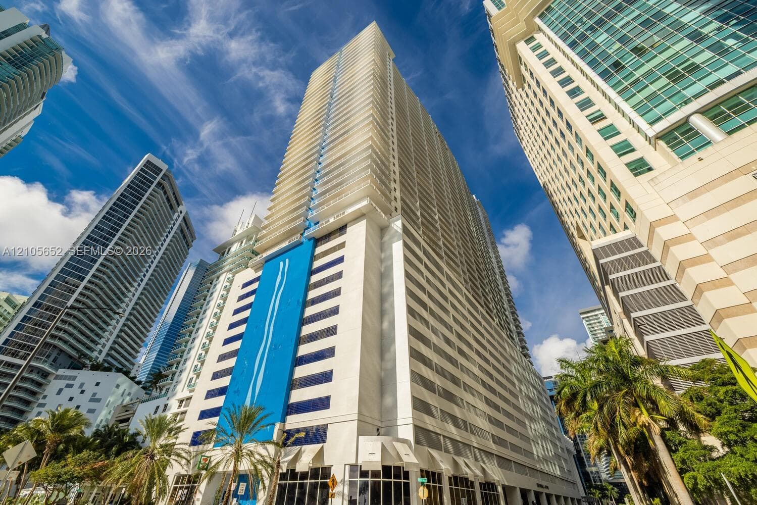 High-rise building exterior featuring a glass-paneled facade, vertical accent wall with blue paneling, integrated balconies, and ground-level glass storefronts