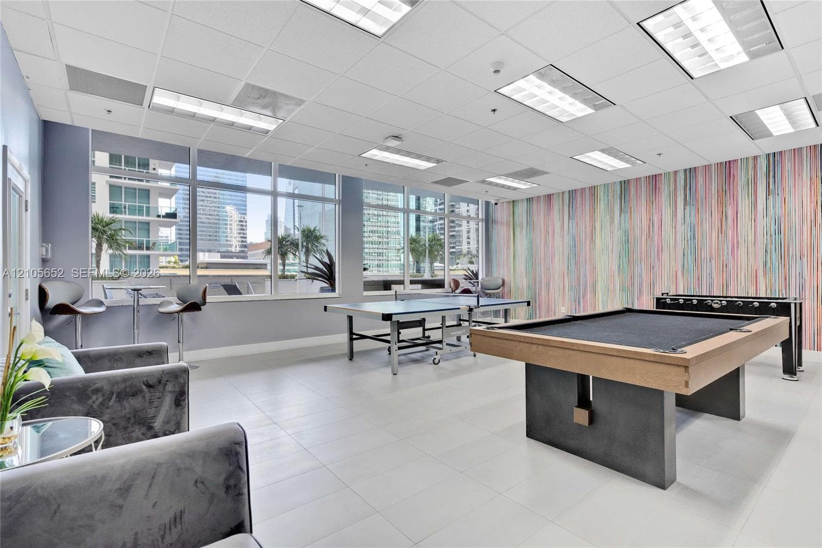 Open-concept recreation area featuring floor-to-ceiling windows, large-format tile flooring, drop ceiling with recessed lighting, and a textured vertical wall finish