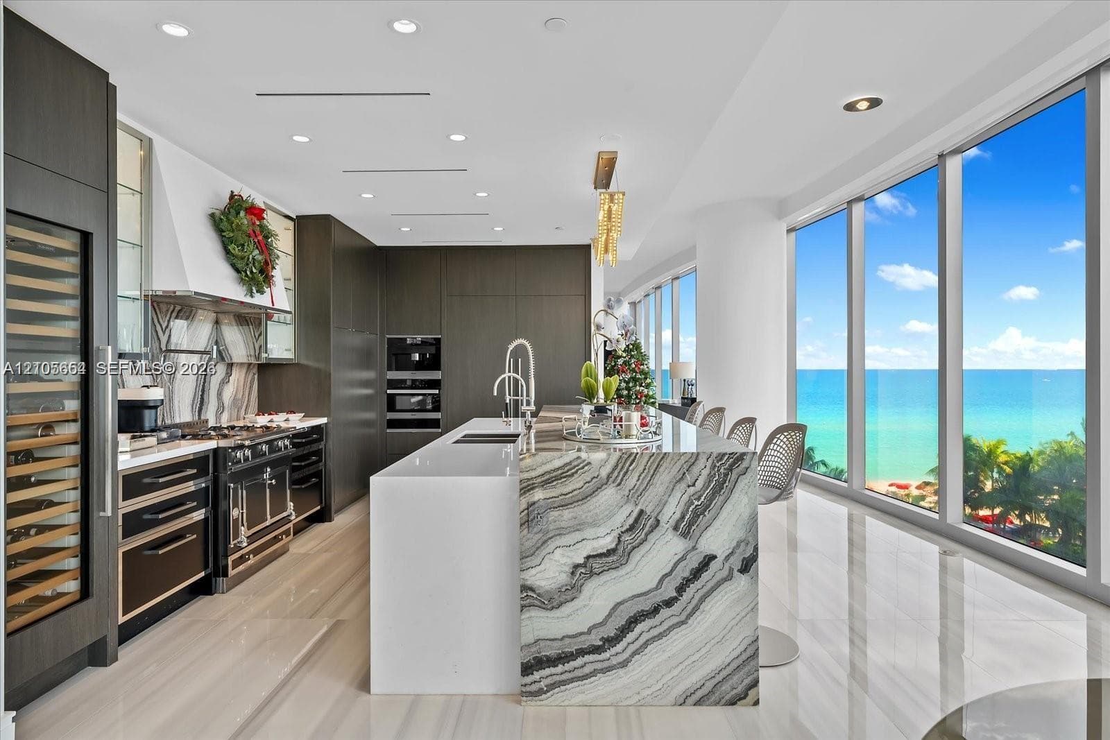Modern kitchen featuring floor-to-ceiling cabinetry, a waterfall-edge island with stone detailing, a multi-burner professional range, integrated wine refrigeration, and expansive floor-to-ceiling windows with ocean scenery