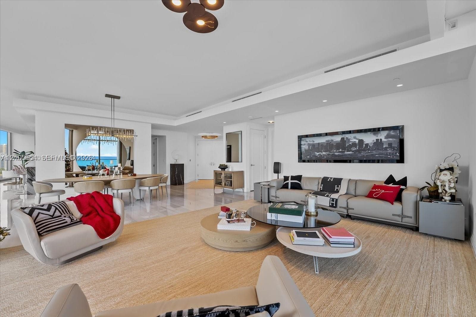 Open-concept interior featuring light-toned stone flooring, recessed ceiling lighting, a modern multi-pendant chandelier, and floor-to-ceiling windows with coastal scenery