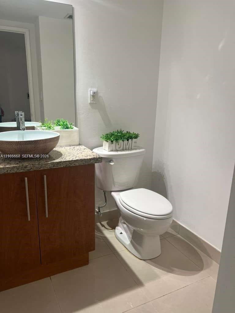 Wood-finish cabinetry, stone-texture countertop, vessel sink with metal faucet, large wall-mounted mirror, and tile flooring