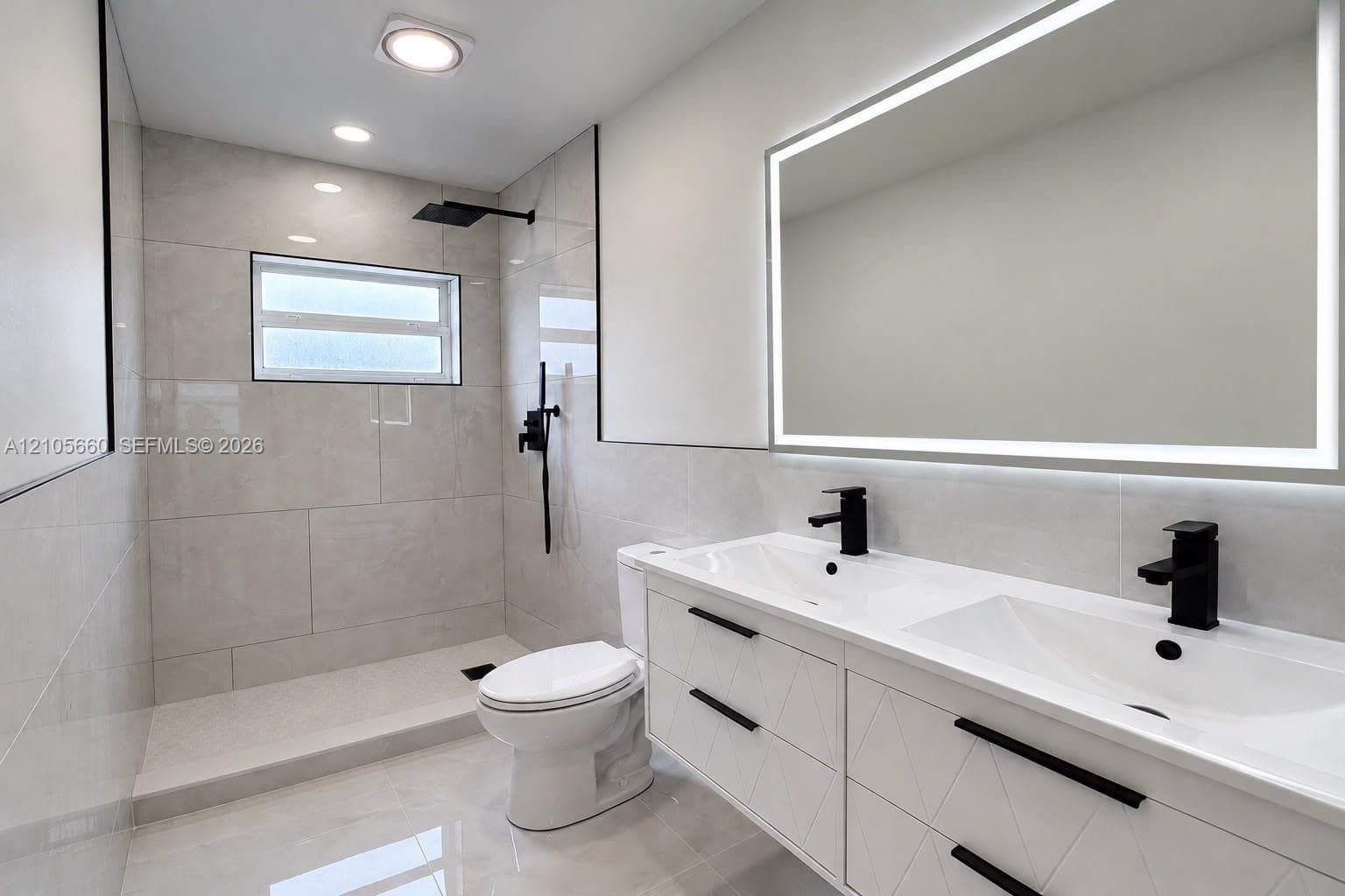 Bathroom featuring a double vanity with white cabinetry, matte black hardware, a rectangular double-basin countertop, a wall-mounted LED mirror, and a walk-in shower with large-format tile surround and matte black rainfall fixture