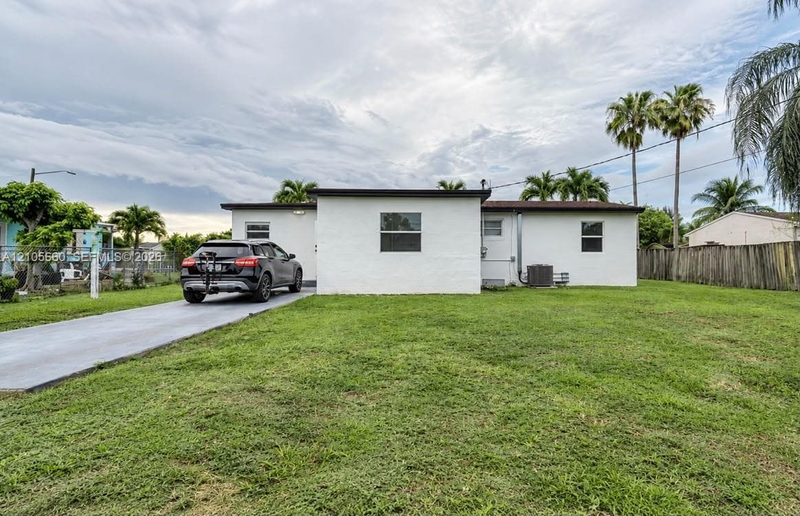 Single-story residence with white masonry exterior, flat roofline, paved concrete driveway, and expansive lawn area enclosed by wooden perimeter fencing