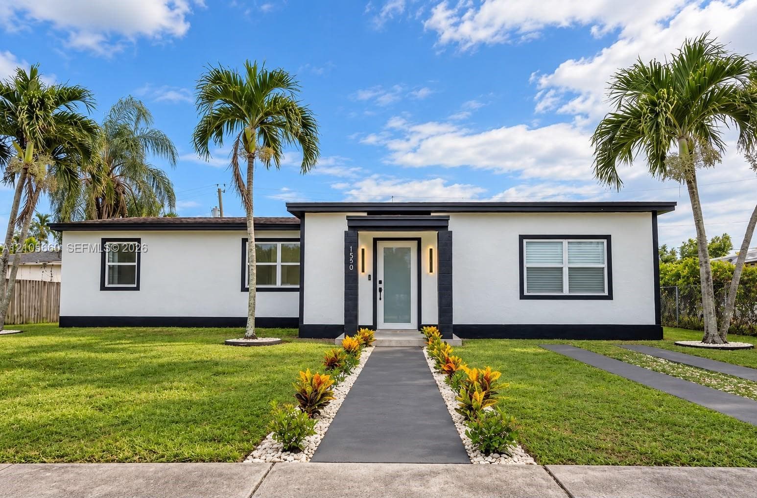 Single-story exterior featuring a high-contrast stucco facade, flat roofline, central recessed entryway with modern vertical lighting, and manicured landscaping with mature palms