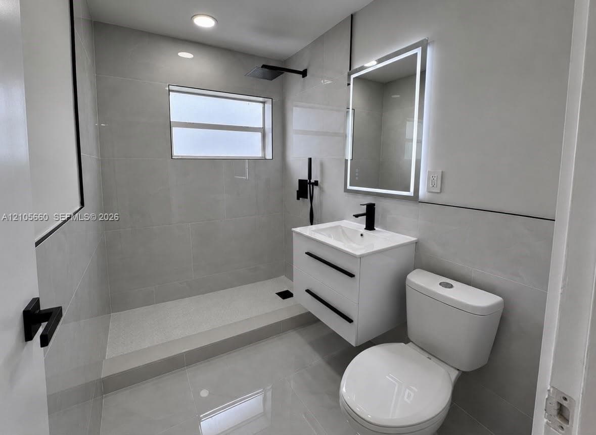 Bathroom featuring large-format gray tile wall and floor surfaces, a floating vanity with a matte black faucet, an LED-backlit mirror, and a walk-in shower with a matte black rainfall showerhead