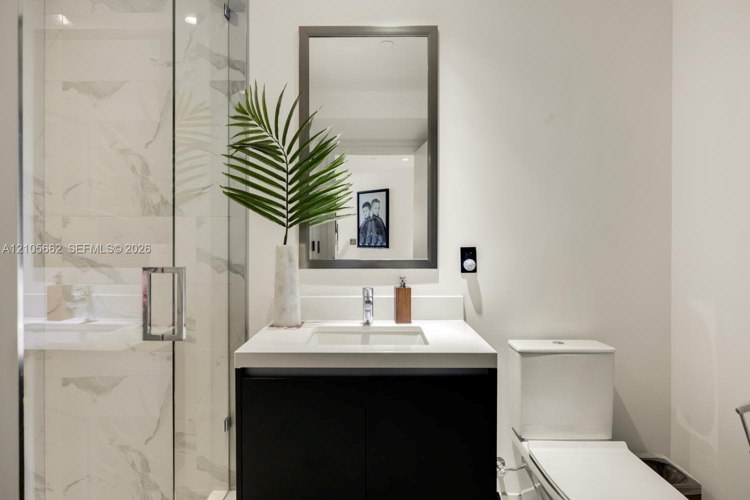 Modern vanity with a countertop basin and cabinet storage, framed wall mirror, glass-enclosed shower with large-format tile surround, and dual-flush toilet