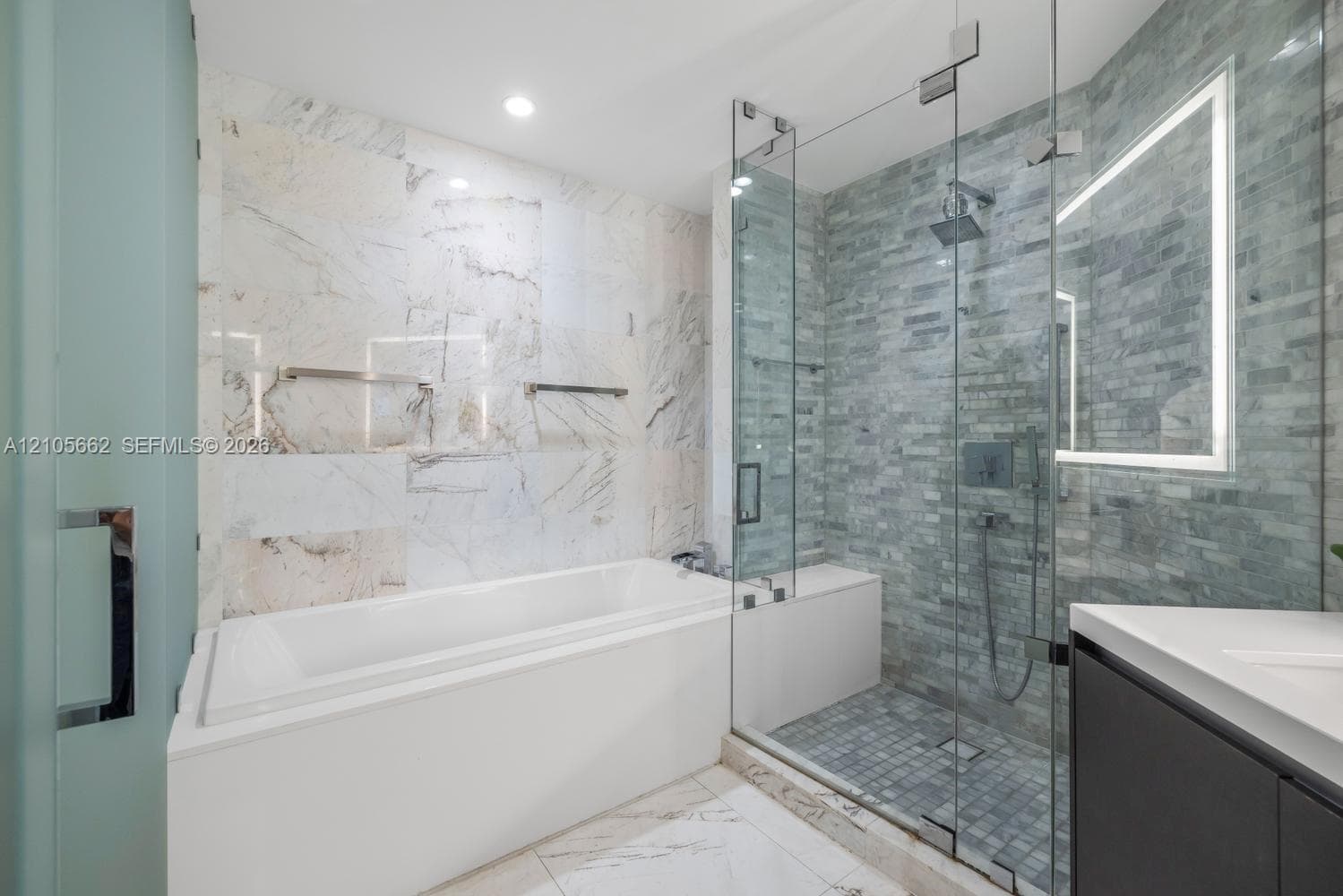 Bathroom featuring a deep soaking tub, walk-in glass-enclosed shower with a built-in bench, textured stone tile wall accents, and an integrated LED-backlit mirror
