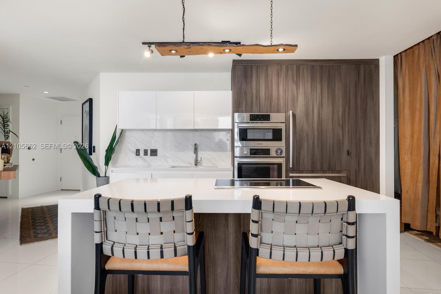 Kitchen featuring a waterfall-edge center island, built-in stainless steel double oven, custom wood-finish cabinetry, natural stone-patterned backsplash, and a rustic live-edge pendant light fixture