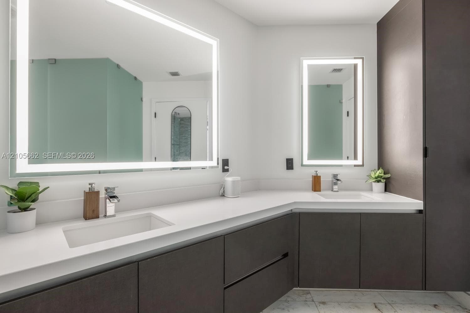 Double vanity featuring dark cabinetry, solid-surface countertops, integrated rectangular sinks, polished chrome faucets, and large backlit mirrors