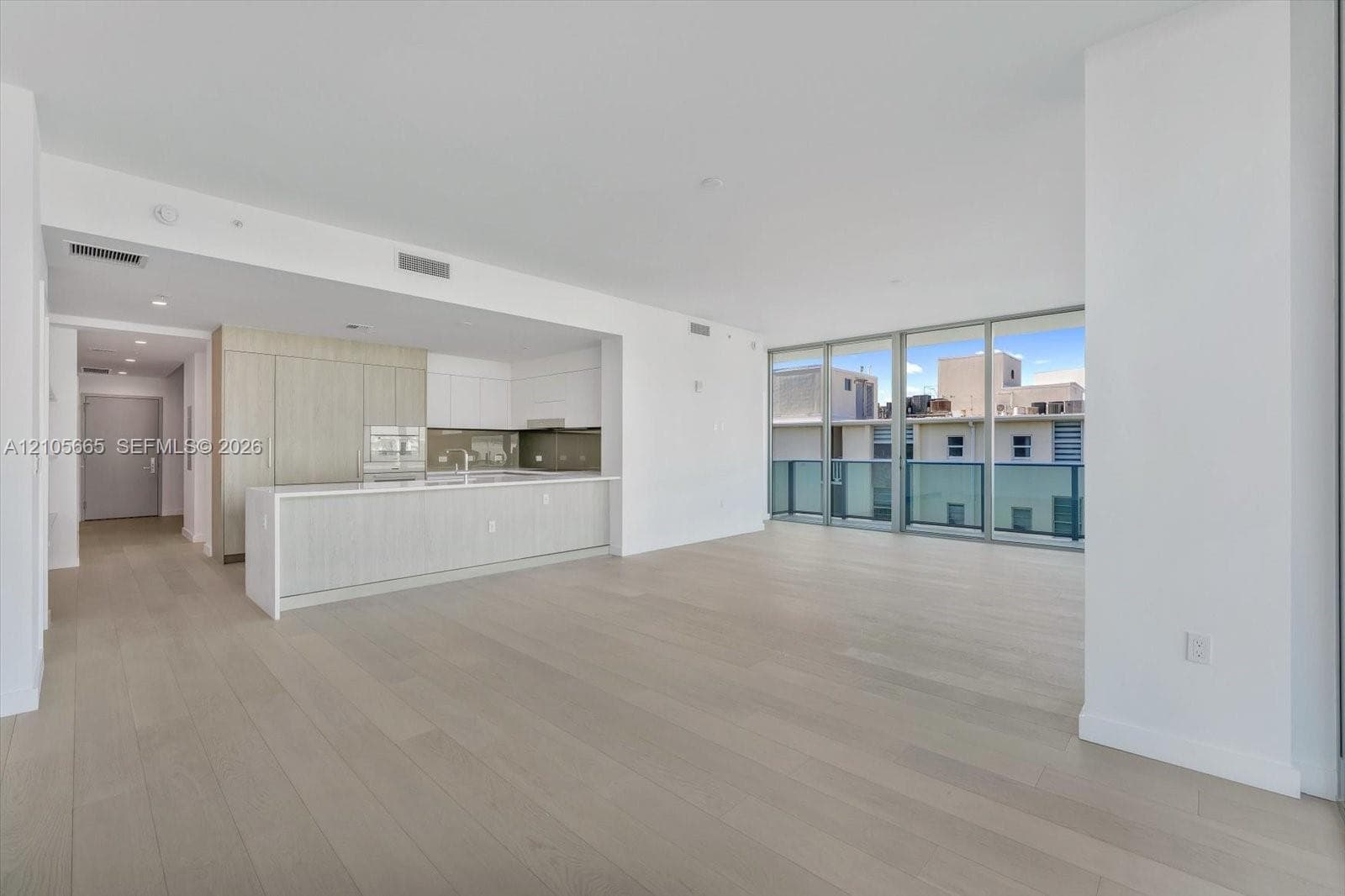 Open-concept interior featuring light-toned wood-finish flooring, floor-to-ceiling glass paneling, and a kitchen equipped with a large island, flat-panel cabinetry, and neutral-toned backsplash