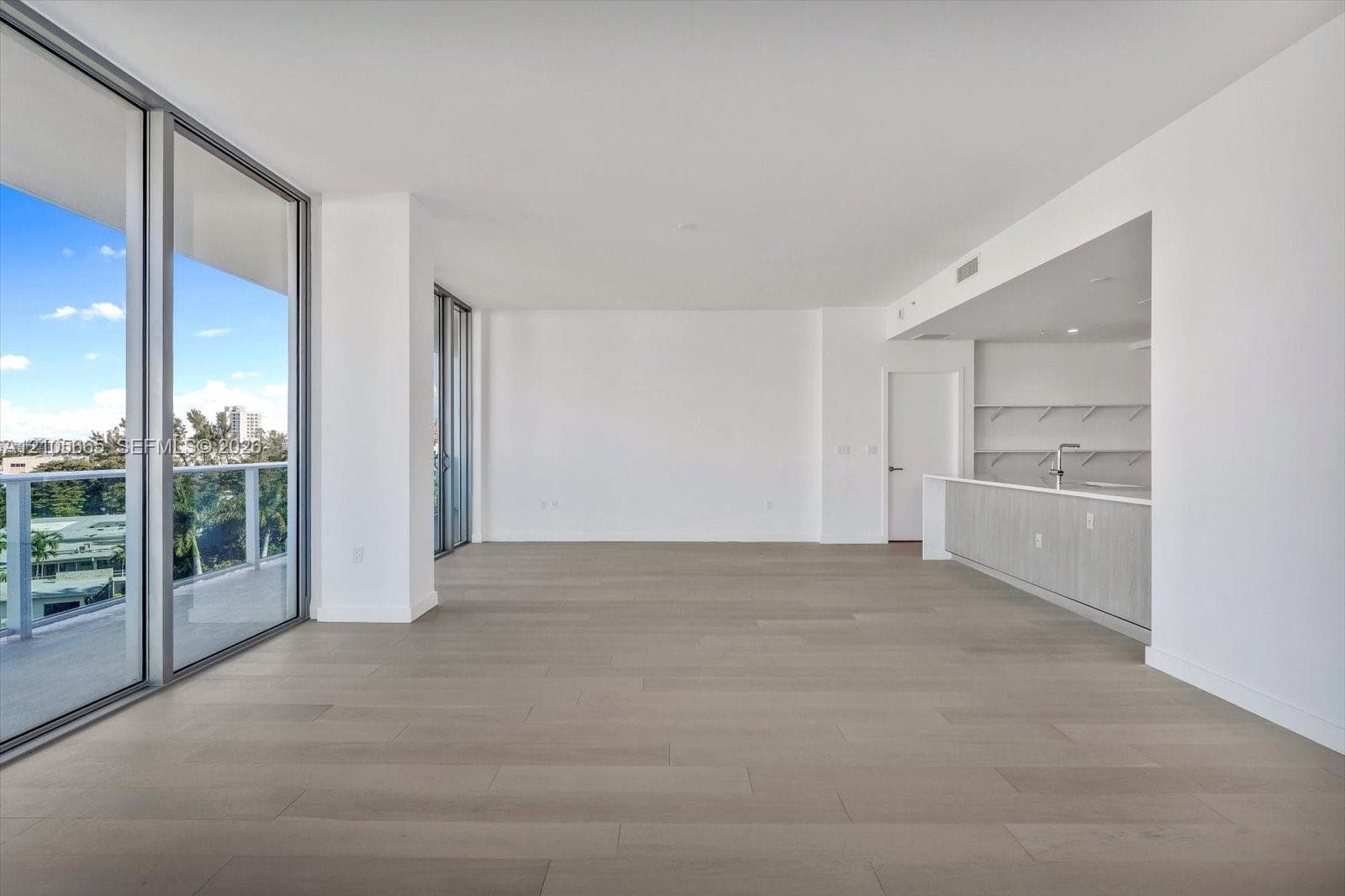 Floor-to-ceiling glass sliding doors, private balcony with metal railing, light-tone wood-finish flooring, and kitchen featuring integrated cabinetry and solid-surface countertops