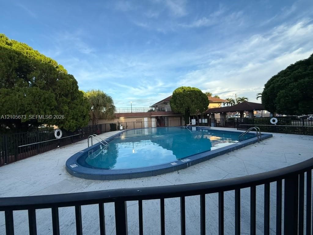 Inground pool with a tiled perimeter, expansive concrete decking, metal safety fencing, and a covered pavilion structure