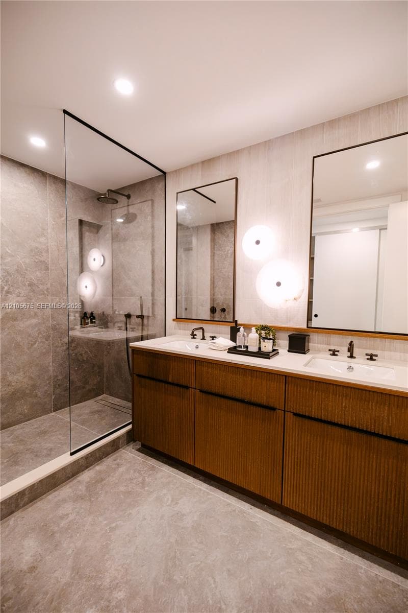 Custom double-sink vanity with fluted wood-finish cabinetry, frameless glass walk-in shower with floor-to-ceiling stone-look tiling, matte black fixtures, and dual wall-mounted vanity mirrors with integrated lighting. Images are for finish purposes only. Please refer to the floor plan for the layout.