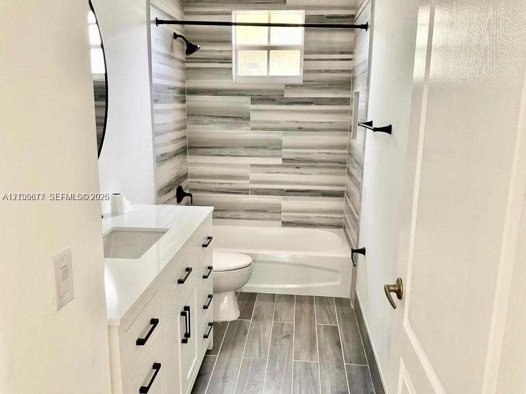 Bathroom featuring a white vanity with a white solid-surface countertop, matte black hardware, horizontal multi-tonal wall tile in the tub enclosure, and wood-finish tile flooring