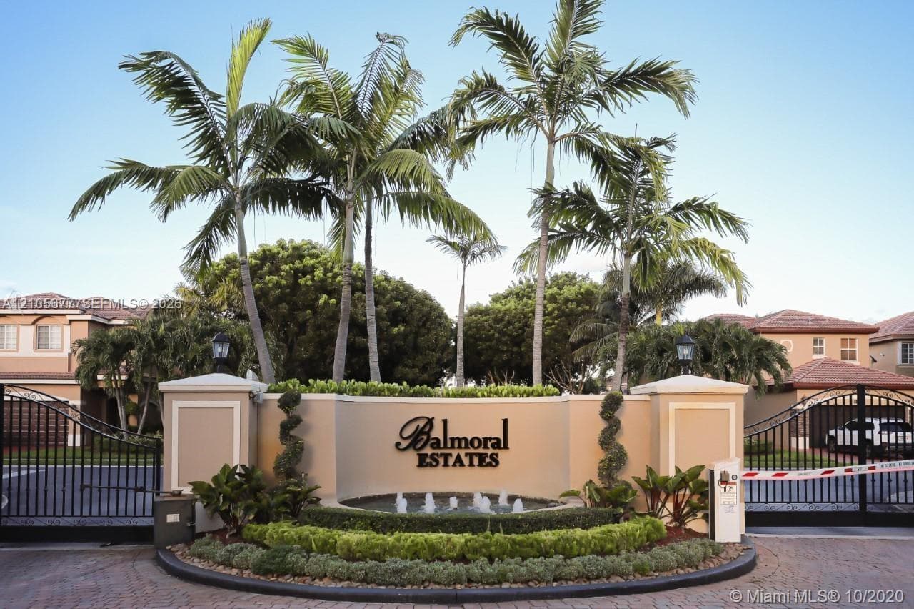 Gated entrance featuring decorative pillars, a tiered fountain, mature palm trees, and structured landscaping