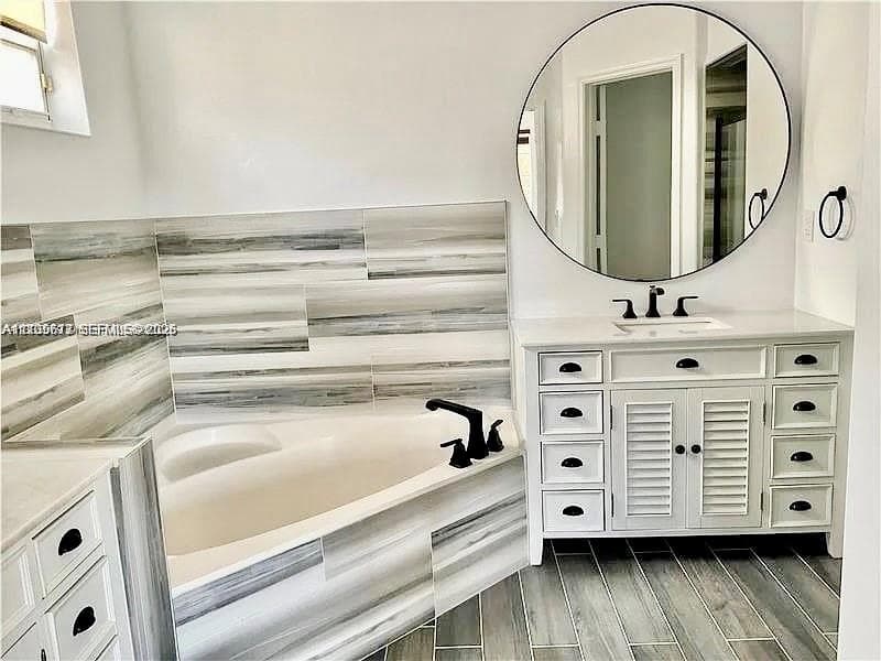Bathroom featuring a white dual-sink vanity with louvered cabinets, a circular mirror, corner soaking tub, horizontal-patterned wall tiling, and wood-finish flooring