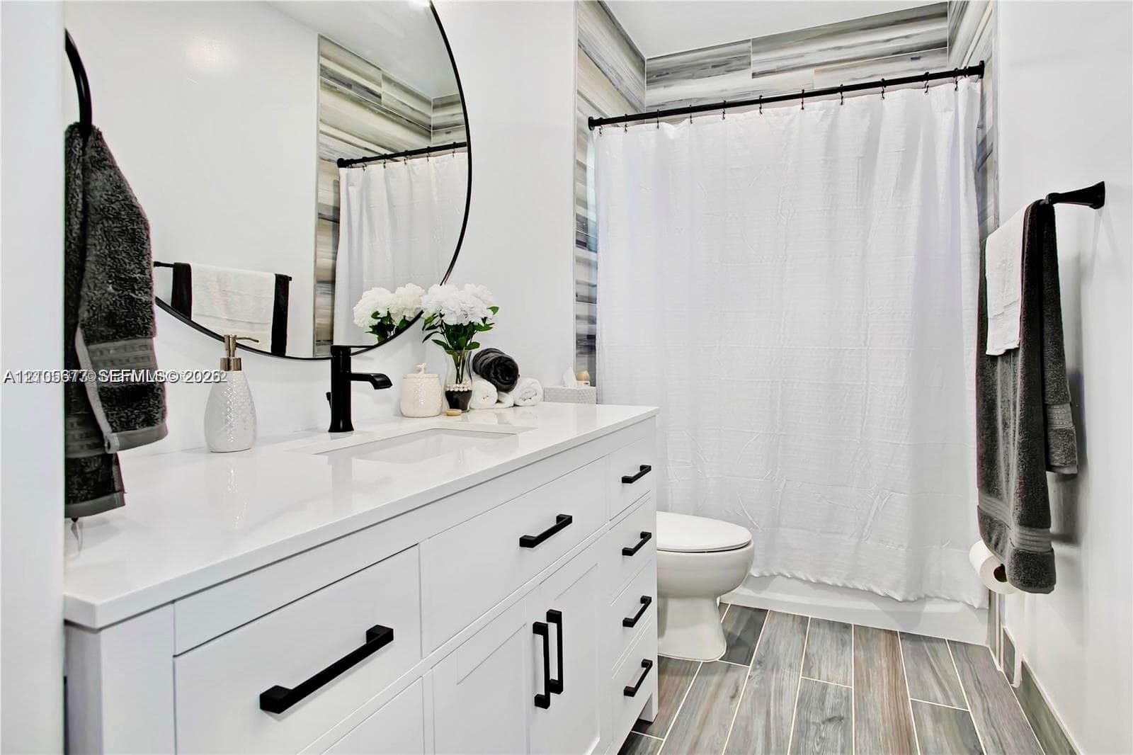 White vanity with solid-surface countertop and matte black hardware, integrated sink, circular mirror, and wood-finish tile flooring