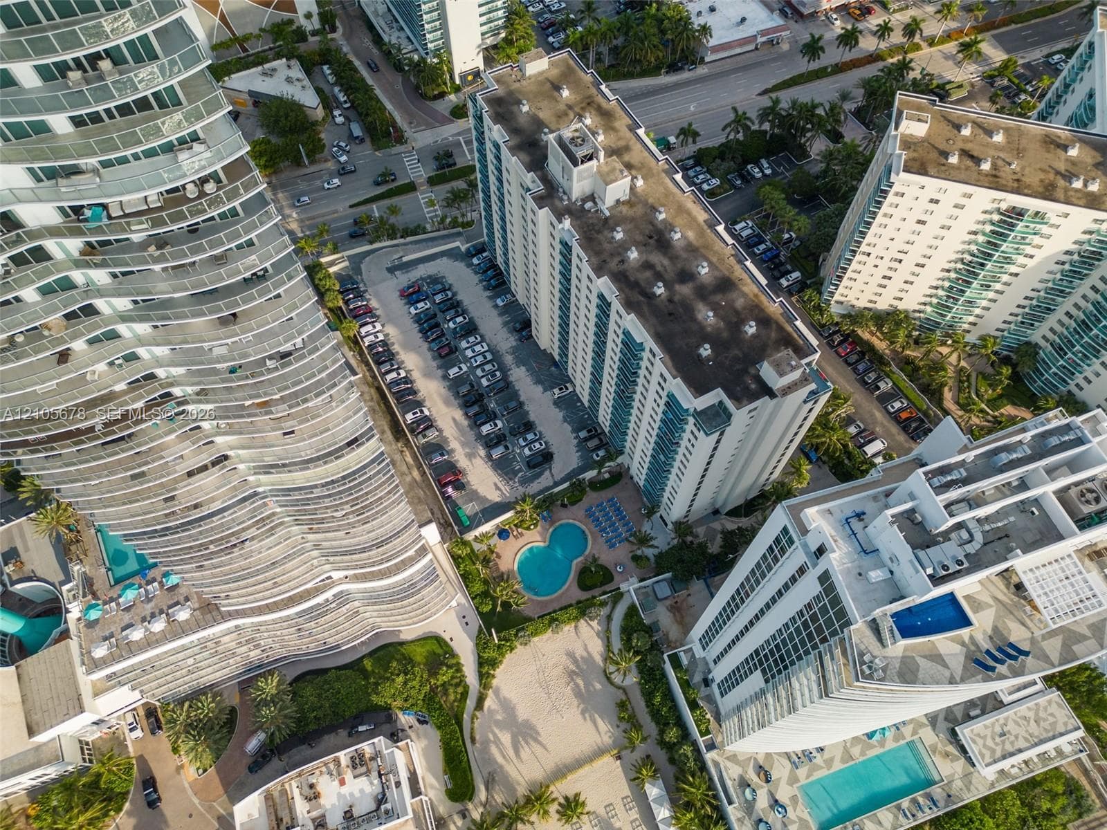 Aerial view showcasing a contemporary residential complex featuring multiple curved-facade towers with balconies, resort-style amenities including swimming pools, manicured landscaping with mature trees, and integrated parking structures