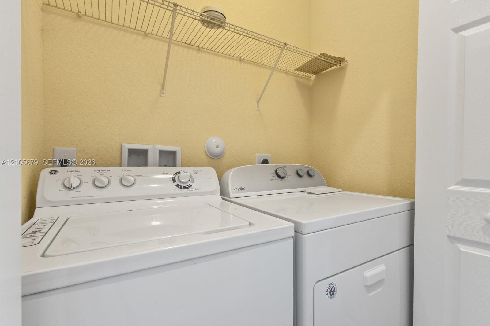 Utility space featuring white top-load washer and dryer units, integrated wall-mounted wire shelving, and recessed electrical and water hookups