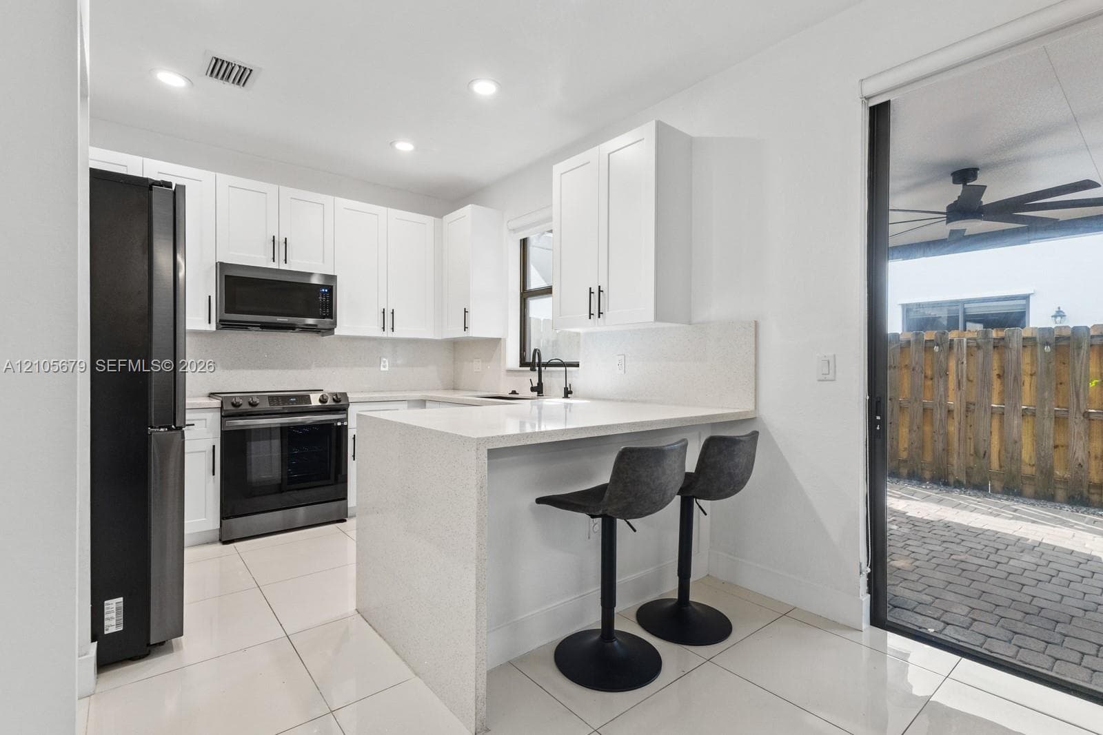 Kitchen featuring white shaker-style cabinetry, quartz countertops with an integrated peninsula, black stainless steel appliances, and large-format tile flooring with access to a paver patio