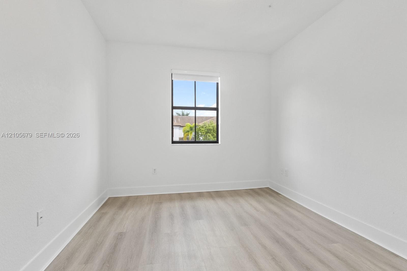 Neutral interior room featuring wood-finish flooring, crisp white walls, and a single window with black grid framing