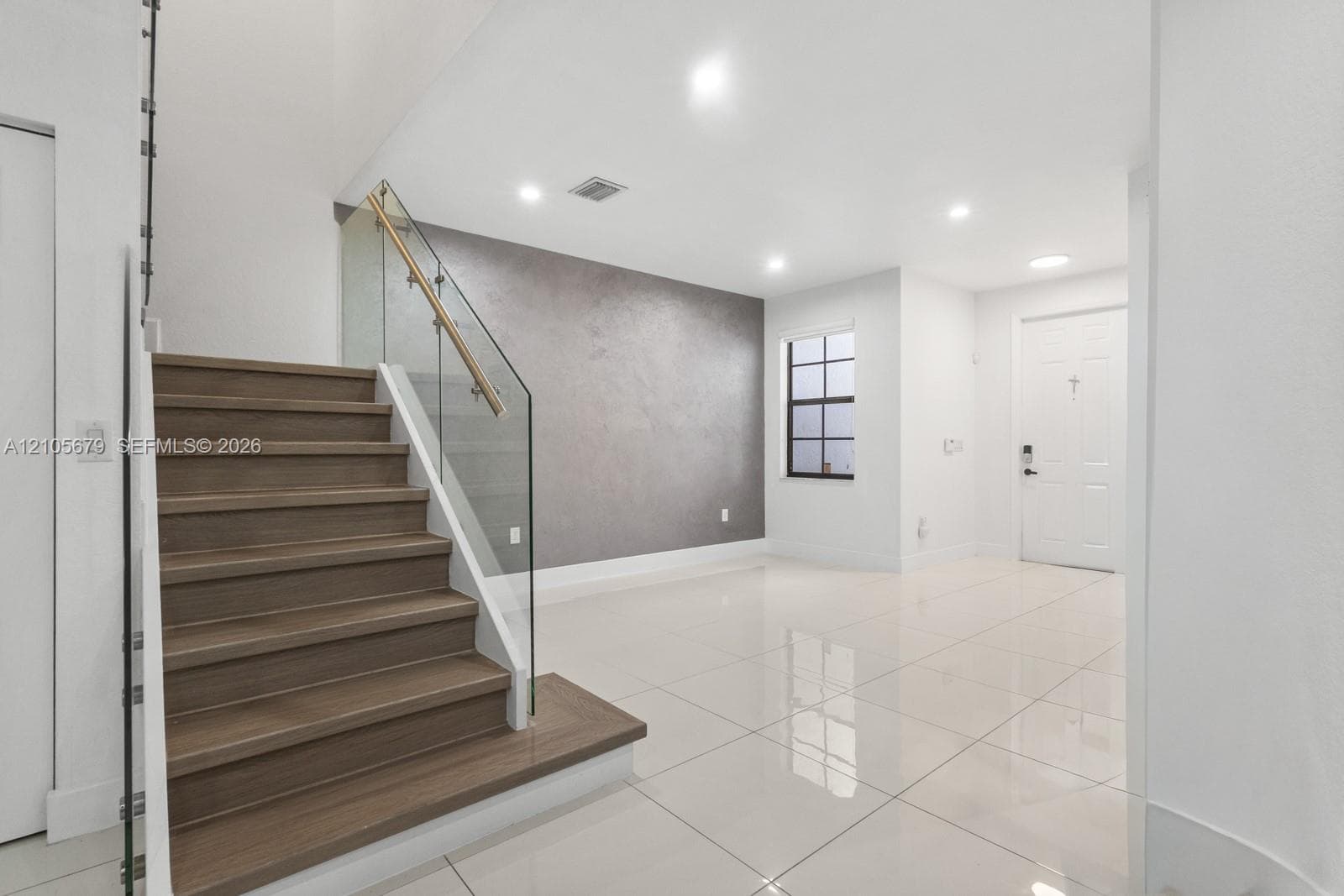 Interior space featuring polished tile flooring, wood-finish stair treads, a glass staircase railing with a metallic handrail, a textured accent wall, and recessed ceiling lighting