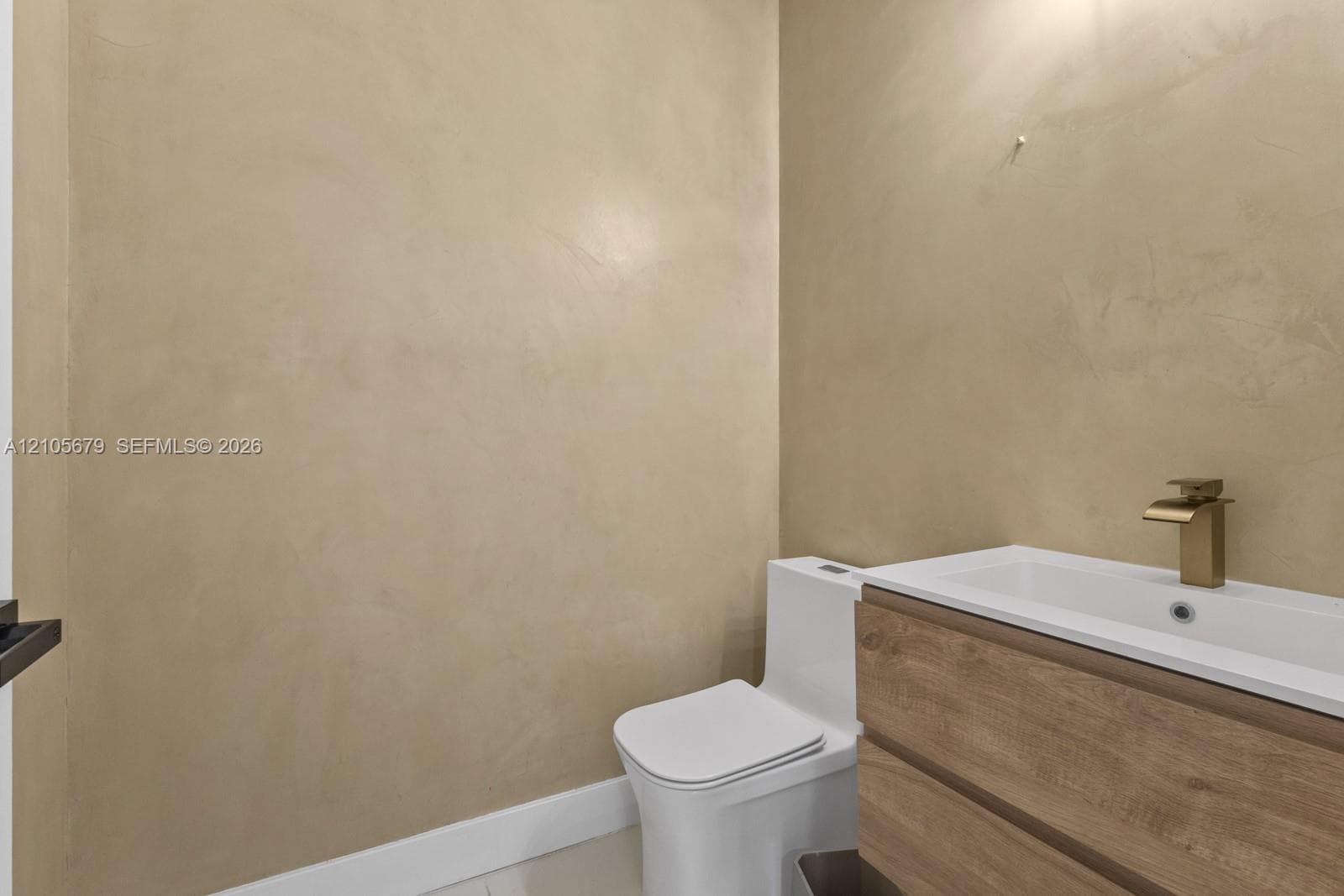 Textured plaster walls paired with a modern wood-finish vanity, integrated rectangular sink, brass-tone faucet, and a one-piece toilet