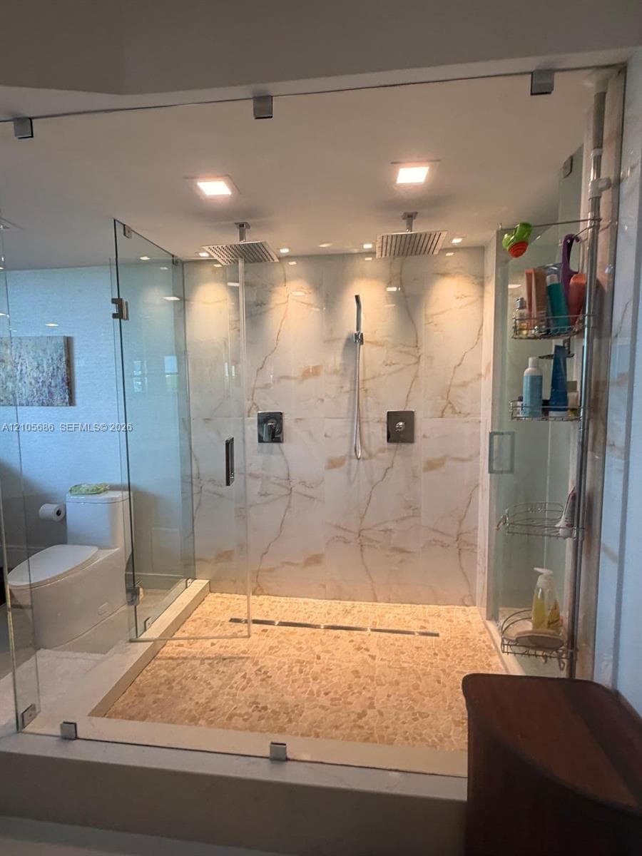 Walk-in shower featuring a frameless glass enclosure, dual rain-style showerheads, hand-held sprayer, large-format stone-look wall tile, and a textured stone mosaic floor