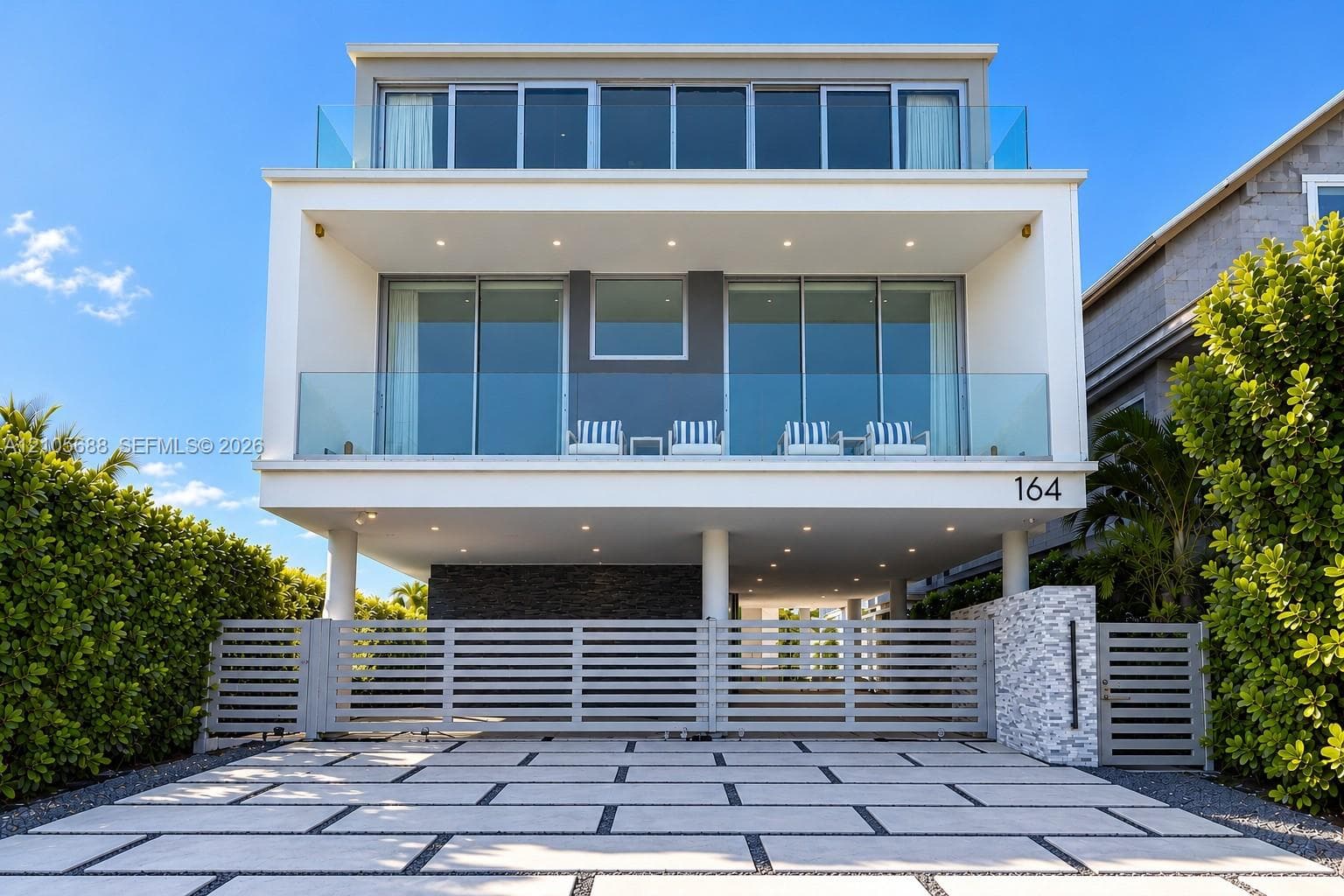 Contemporary architectural exterior featuring stacked-stone masonry, multi-level glass balconies with frameless railings, oversized floor-to-ceiling sliding glass panels, and a custom paver-grid driveway with horizontal slat gate
