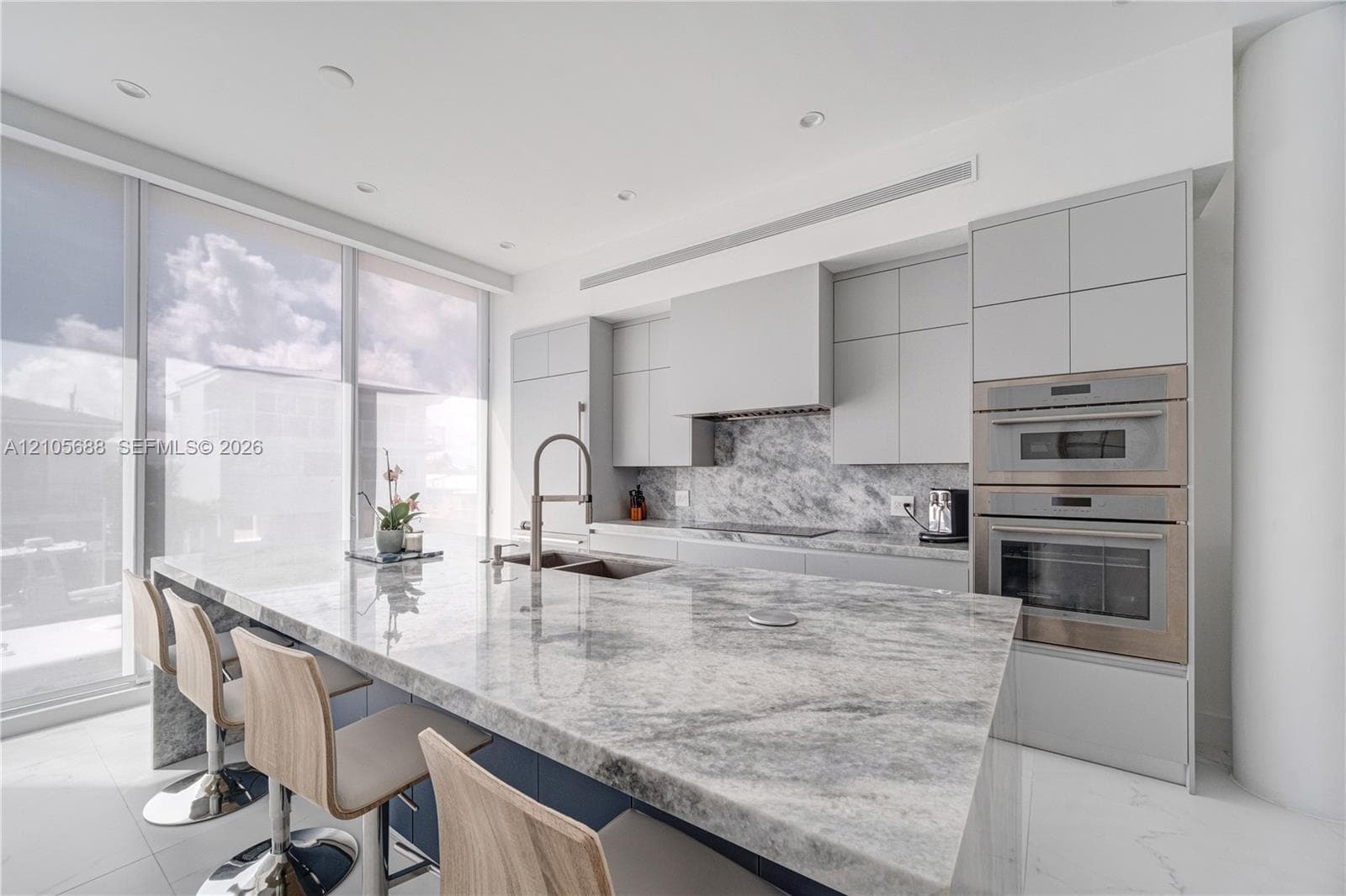 Modern kitchen featuring floor-to-ceiling windows, a large stone-surfaced waterfall island, matching stone slab backsplash, flat-panel cabinetry, and integrated stainless steel double wall ovens
