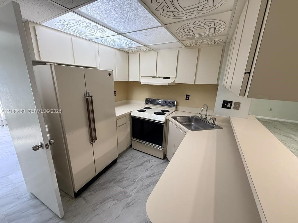 Kitchen featuring neutral cabinetry, solid-surface countertops, a stainless steel sink with a high-arch faucet, an electric range, and patterned drop ceiling tiles