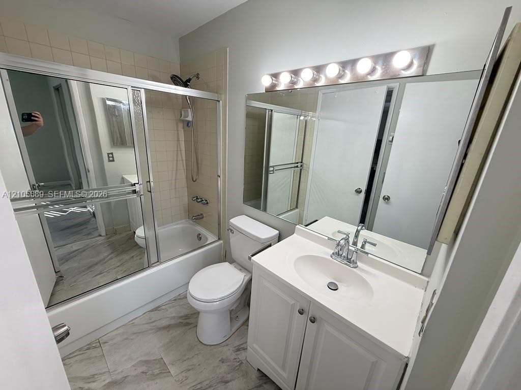 Bathroom featuring a vanity with a solid-surface countertop, full-width vanity mirror, tile-surround tub-shower combination with sliding glass doors, and light-tone stone-finish flooring
